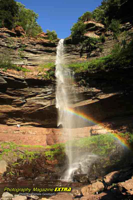 Kaaterskill falls from the top to the bottom, with a big rainbow with two people behind the falls.