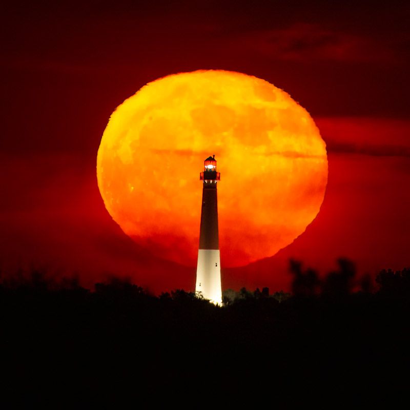 A lighthouse with a very large moon behind it. or could be a sun.
