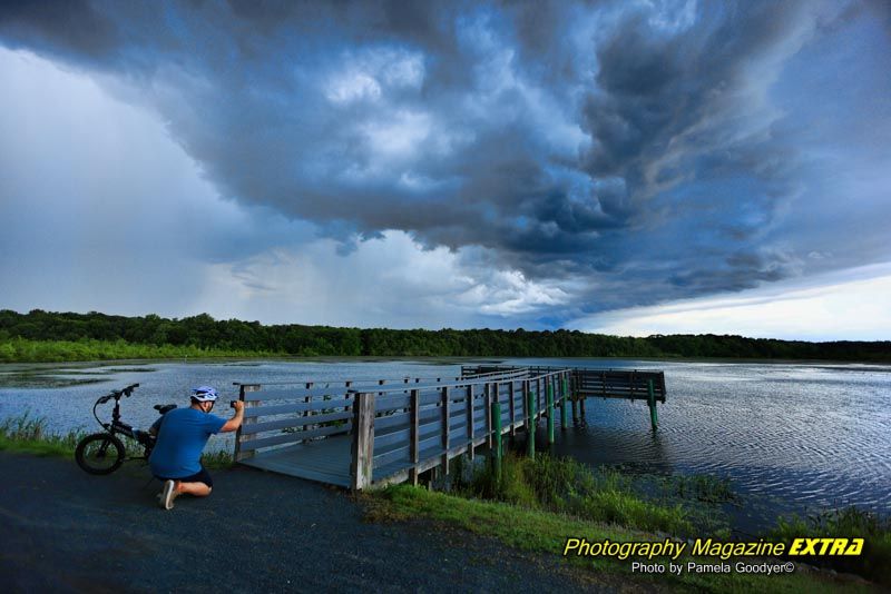 Helmet a lake with very dark, cloudy skies and a man taking a picture of the dock. Next to his bicycle.